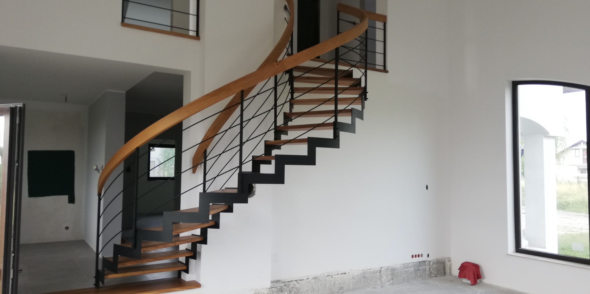 Spiralne schody drewniane z czarnymi metalowymi elementami w jasnym wnętrzu z dużym oknem, widoczne niedokończone wykończenie pod ścianą.