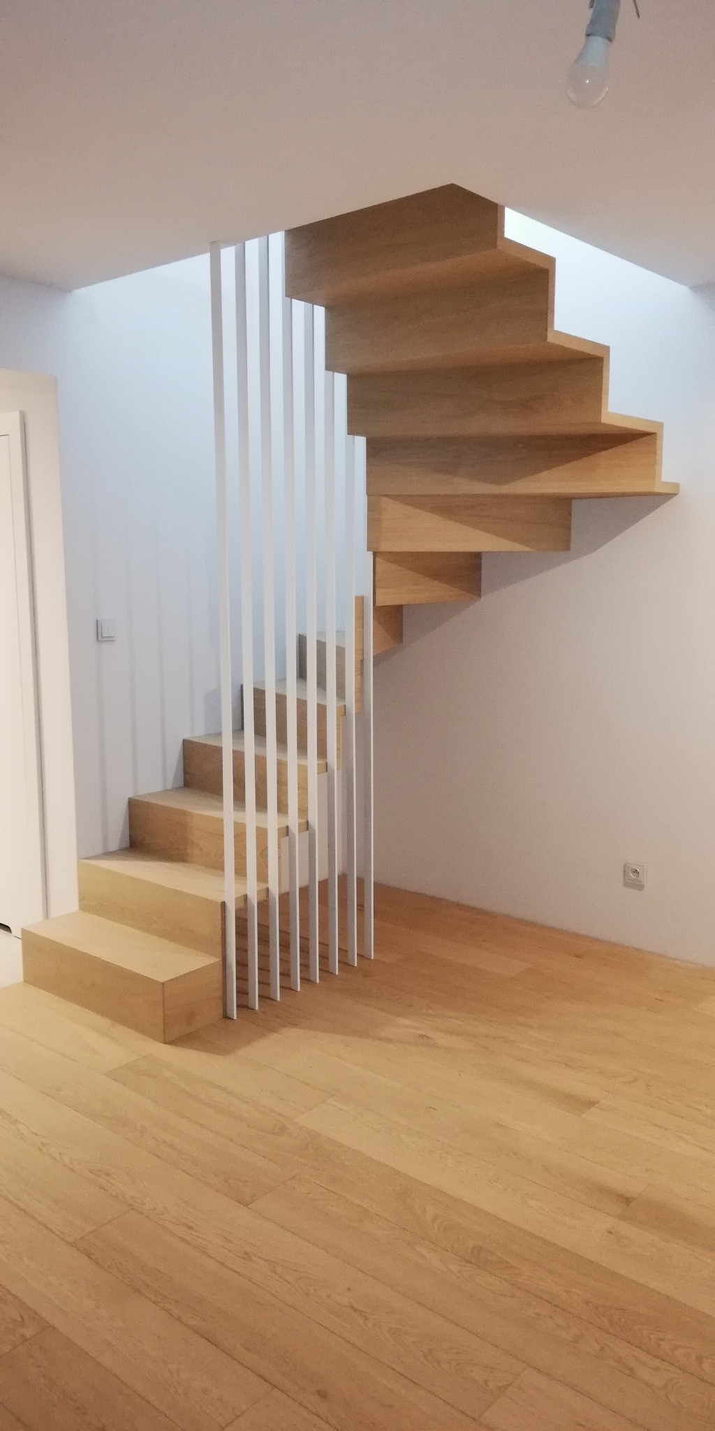 Nowoczesne, drewniane schody dywanowe z białymi, pionowymi balustradami w minimalistycznym wnętrzu z parkietem.