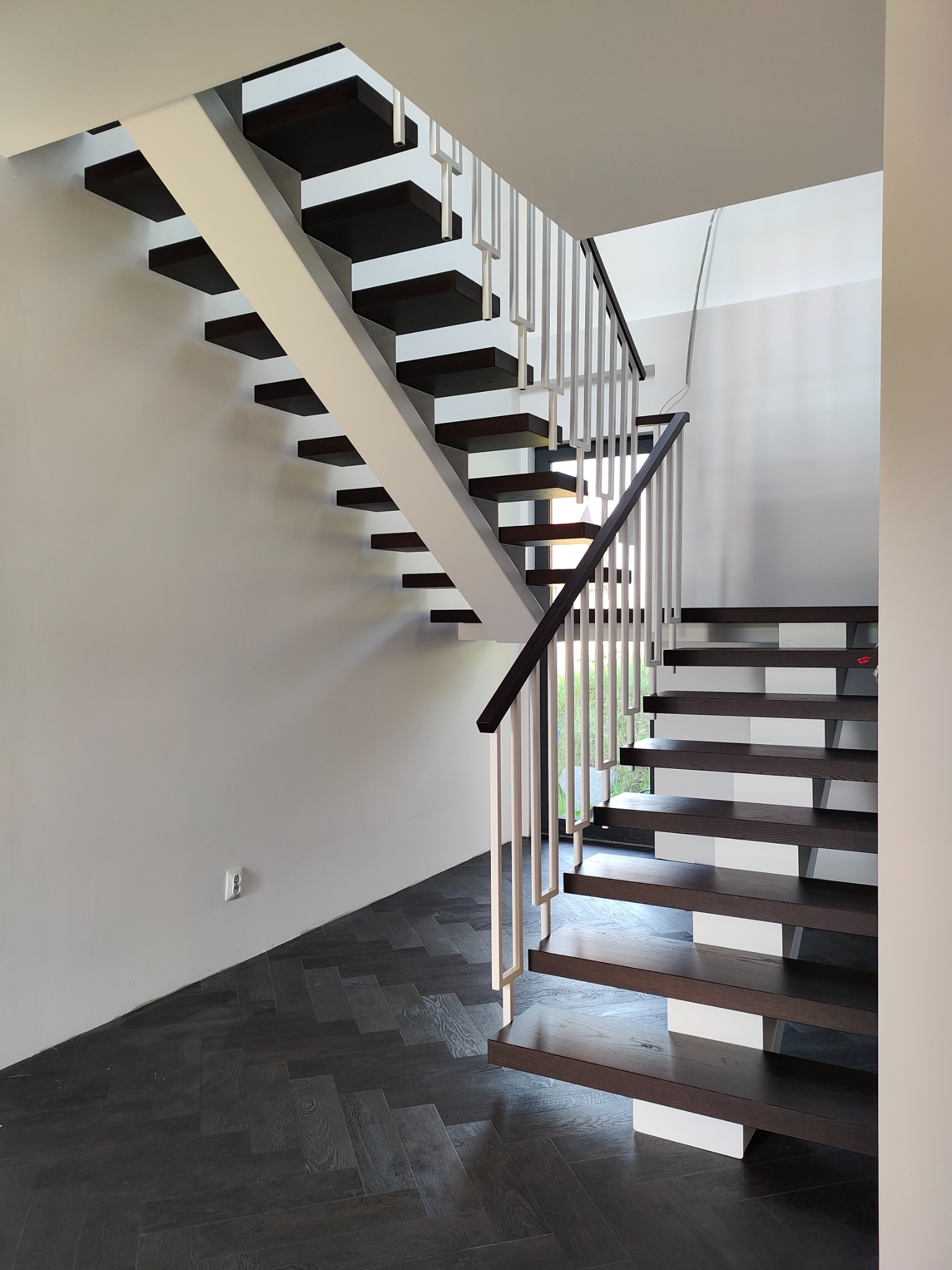 Nowoczesne schody z ciemnego drewna z białymi wspornikami i balustradą, parkiet w jodełkę na podłodze, minimalistyczne wnętrze.