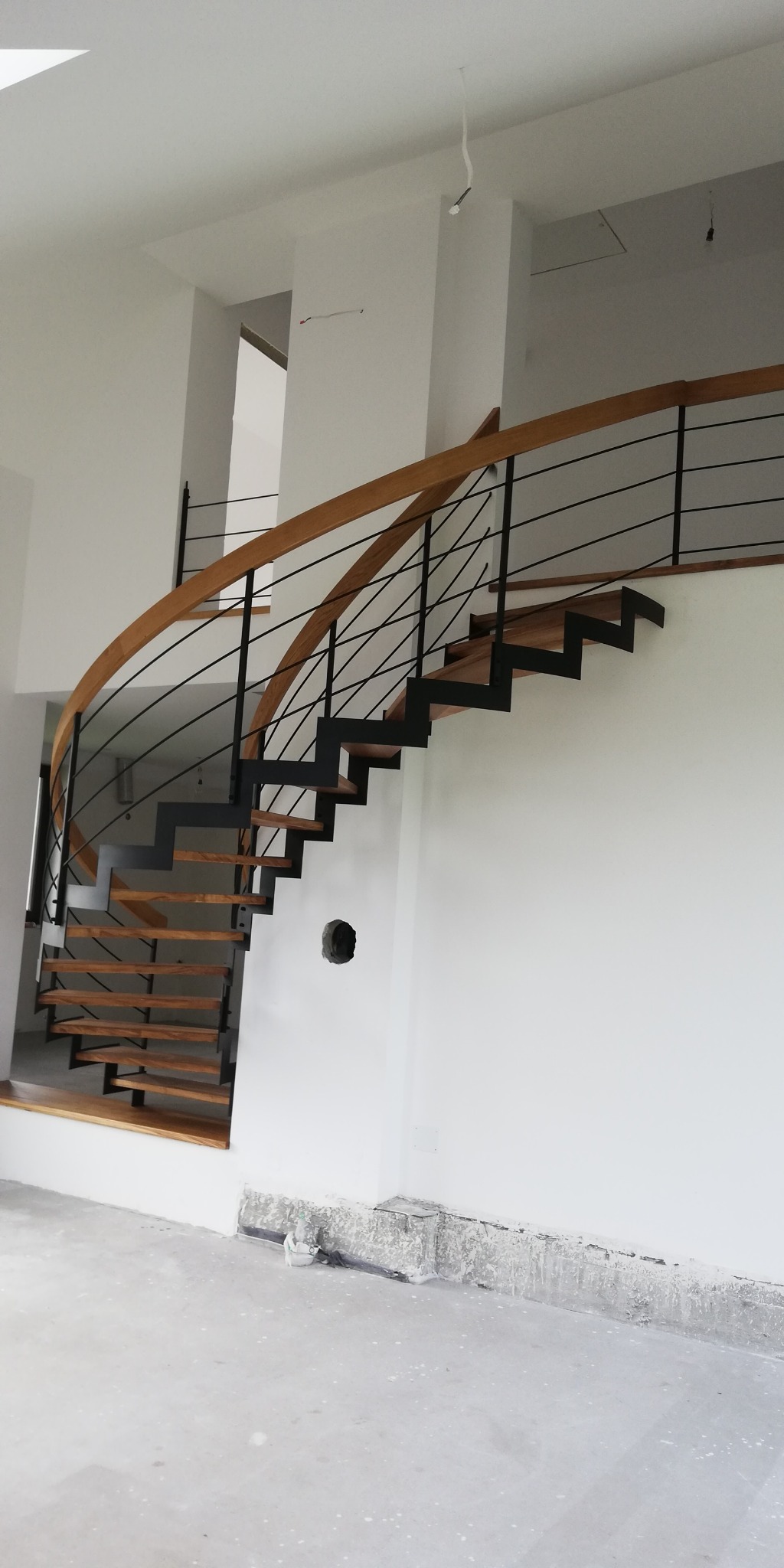 Spiralne schody z drewnianymi stopniami i czarną metalową balustradą w surowym wnętrzu, widoczne przygotowania do wykończenia podłogi.