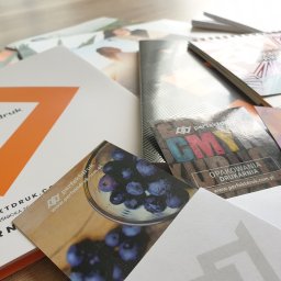 PERFEKTDRUK Joanna Zięba - Stos różnorodnych materiałów reklamowych i poligraficznych z logo 'perfektdruk', w tym wizytówki z motywem owoców, kalendarz spiralny z geometrycznym wzorem i ulotki z abstrakcyjnymi grafikami...