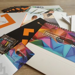 PERFEKTDRUK Joanna Zięba - Stos różnorodnych materiałów drukowanych, w tym kalendarz spiralny z geometrycznym wzorem, wizytówki z logo firmy, ulotki i katalogi, prezentujących usługi drukarni.