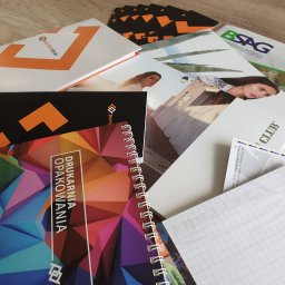 PERFEKTDRUK Joanna Zięba - Stos materiałów drukowanych, w tym notes ze spiralą, ulotki i wizytówki z logo firmy poligraficznej, położone na drewnianym stole.