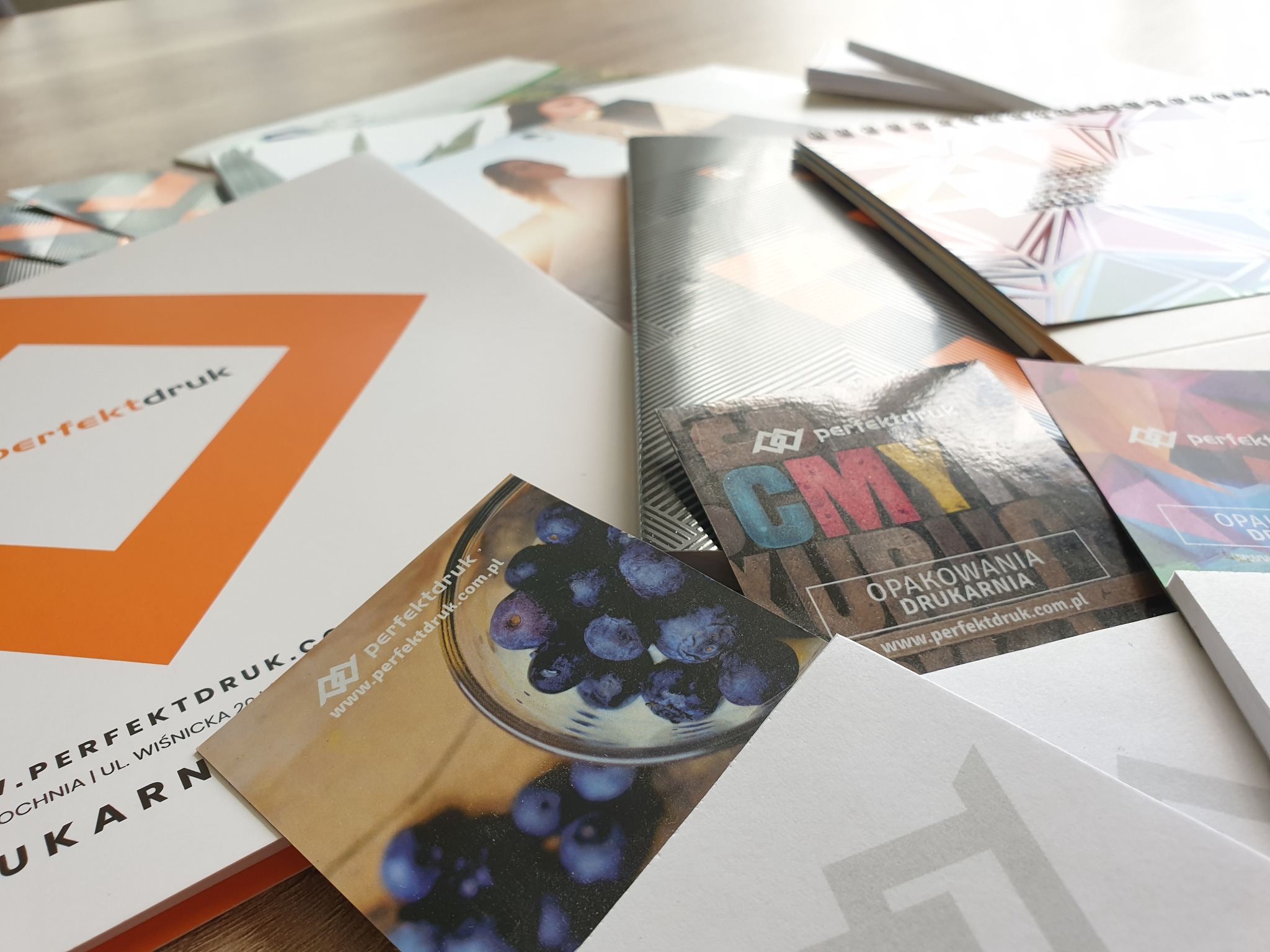 Stos różnorodnych materiałów reklamowych i poligraficznych z logo 'perfektdruk', w tym wizytówki z motywem owoców, kalendarz spiralny z geometrycznym wzorem i ulotki z abstrakcyjnymi grafikami...
