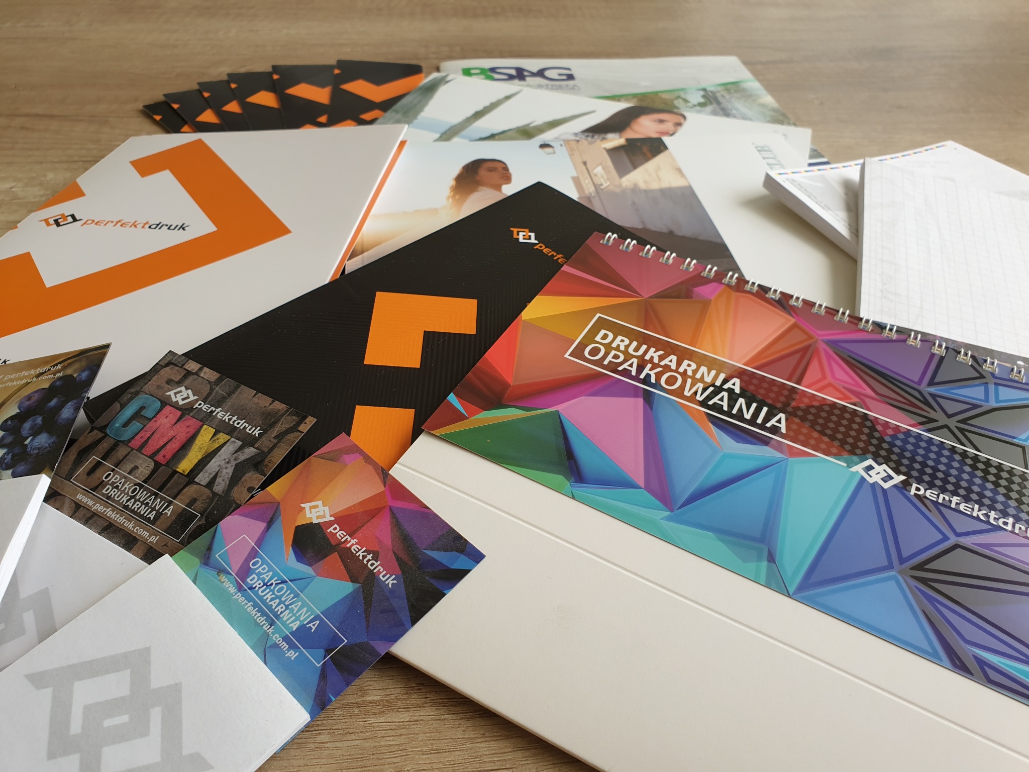 Stos różnorodnych materiałów drukowanych, w tym kalendarz spiralny z geometrycznym wzorem, wizytówki z logo firmy, ulotki i katalogi, prezentujących usługi drukarni.