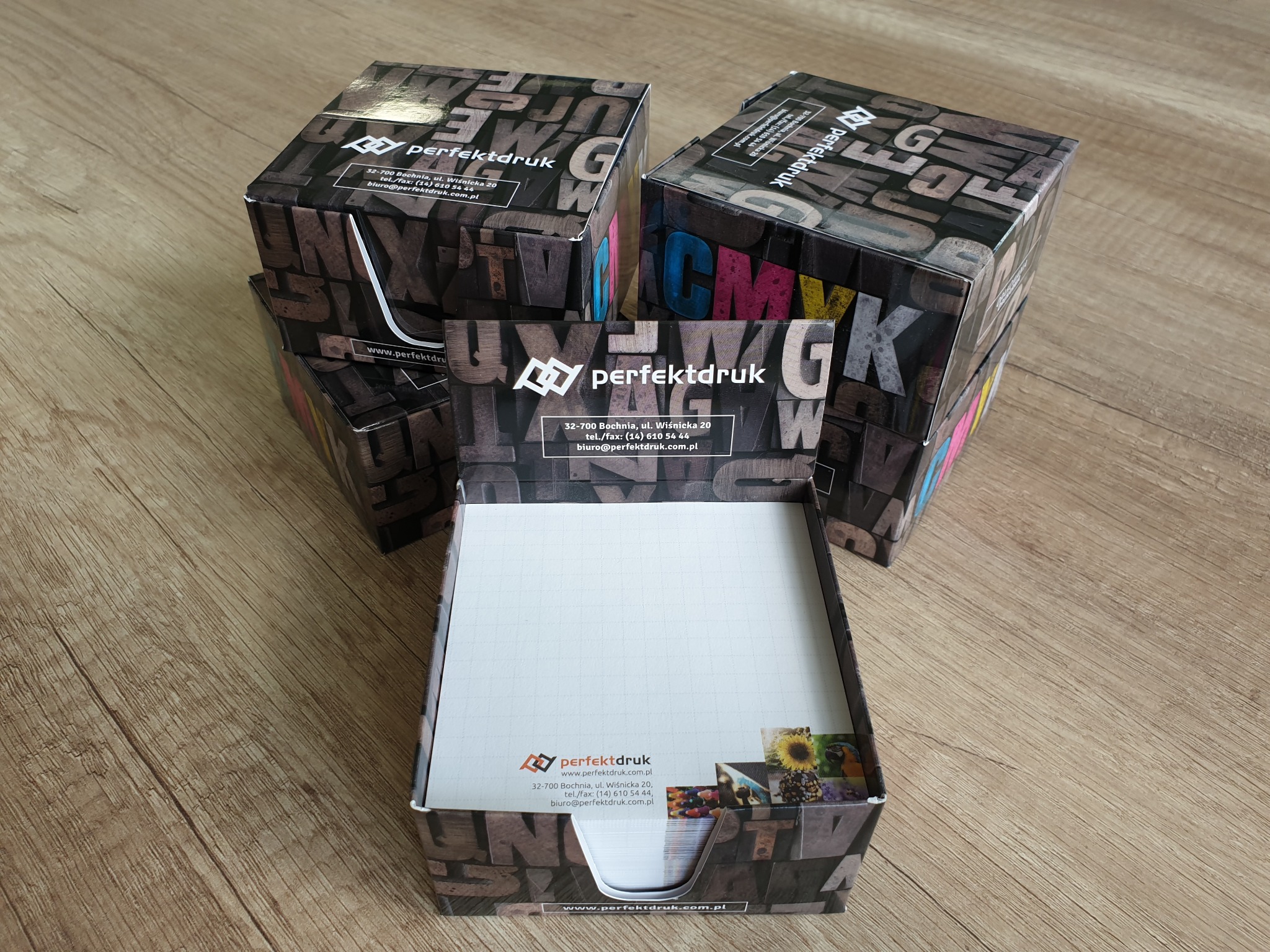 Stos pudełek na notatki z nadrukiem typograficznym i logo firmy perfektdruk, ułożonych na drewnianej powierzchni.