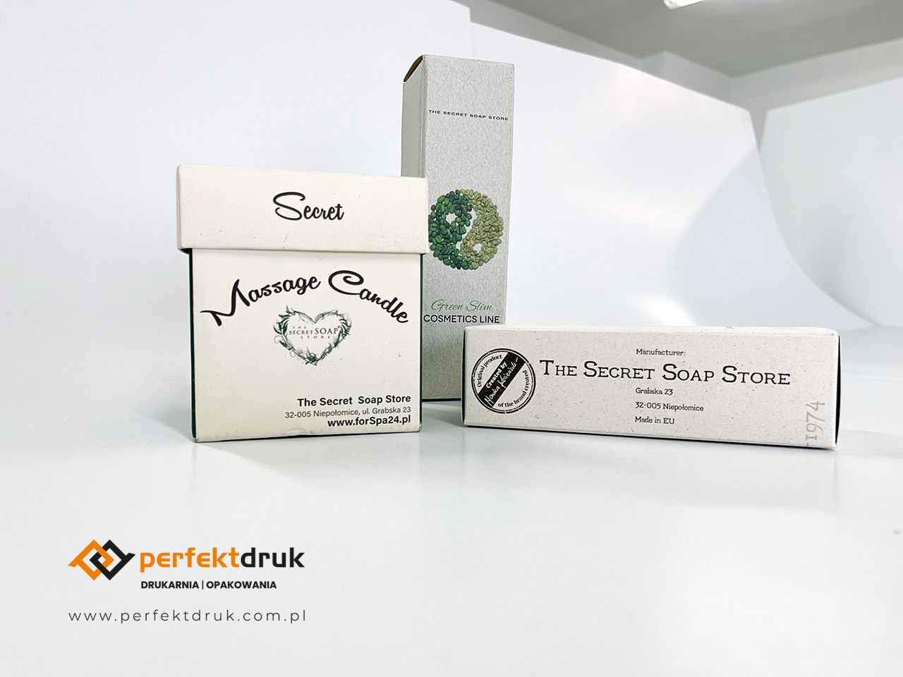 Trzy pudełka kosmetyczne w różnych rozmiarach i kształtach, prezentujące markę The Secret Soap Store, na białym tle z widocznym logo drukarni Perfekt Druk w dolnej części kadru.