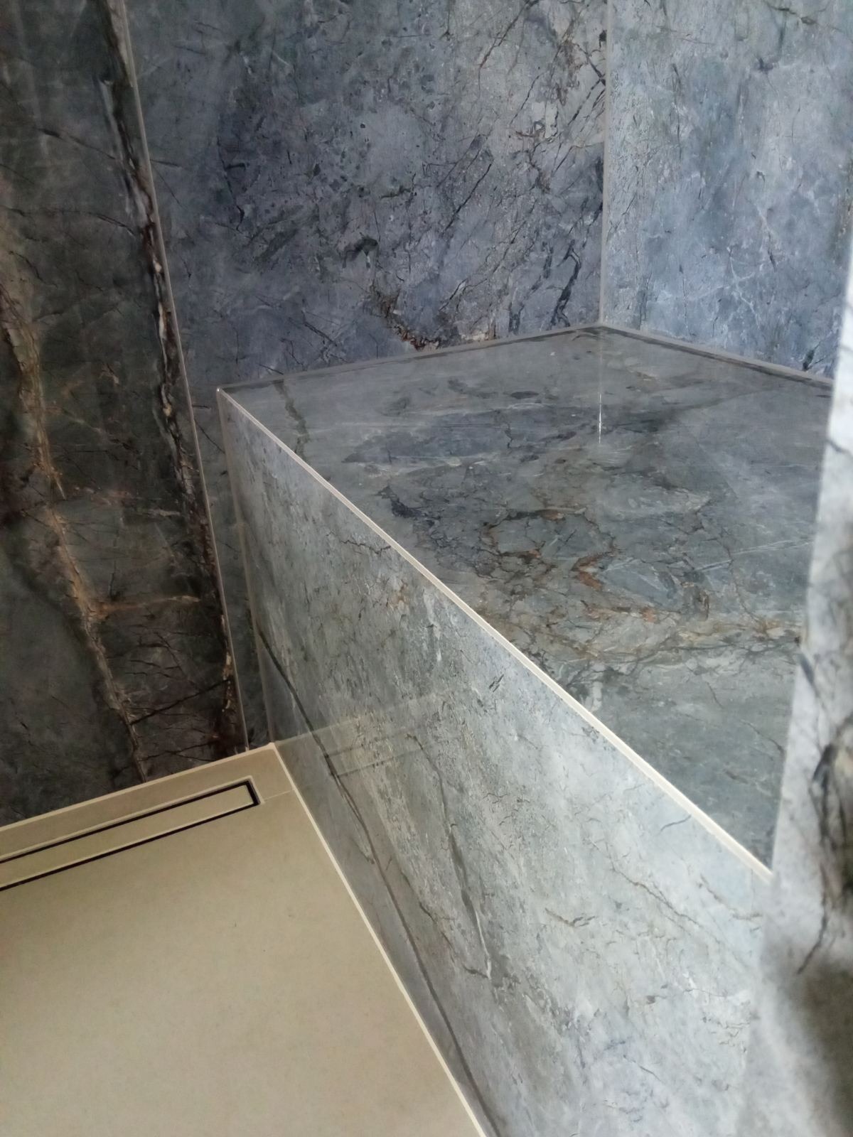 Wykończony narożnik prysznica z szarymi płytkami imitującymi marmur, widoczny odpływ liniowy i półka narożna z tego samego materiału.
