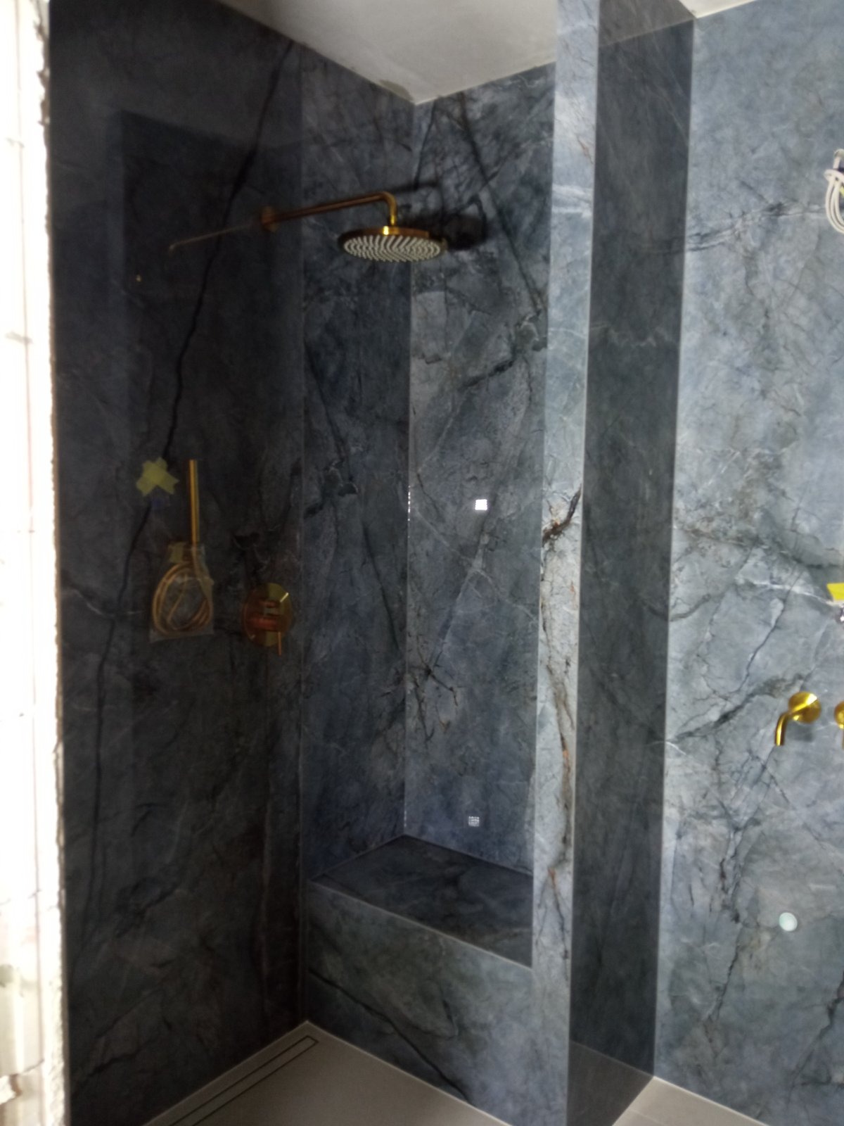 Kabina prysznicowa wykończona płytami imitującymi szary marmur z złotą armaturą, wbudowane siedzisko.