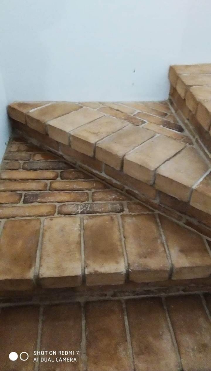 Schody wyłożone ceglanymi płytkami, widoczne spoiny i niedoskonałości faktury płytek, ściana pomalowana na biało w tle.