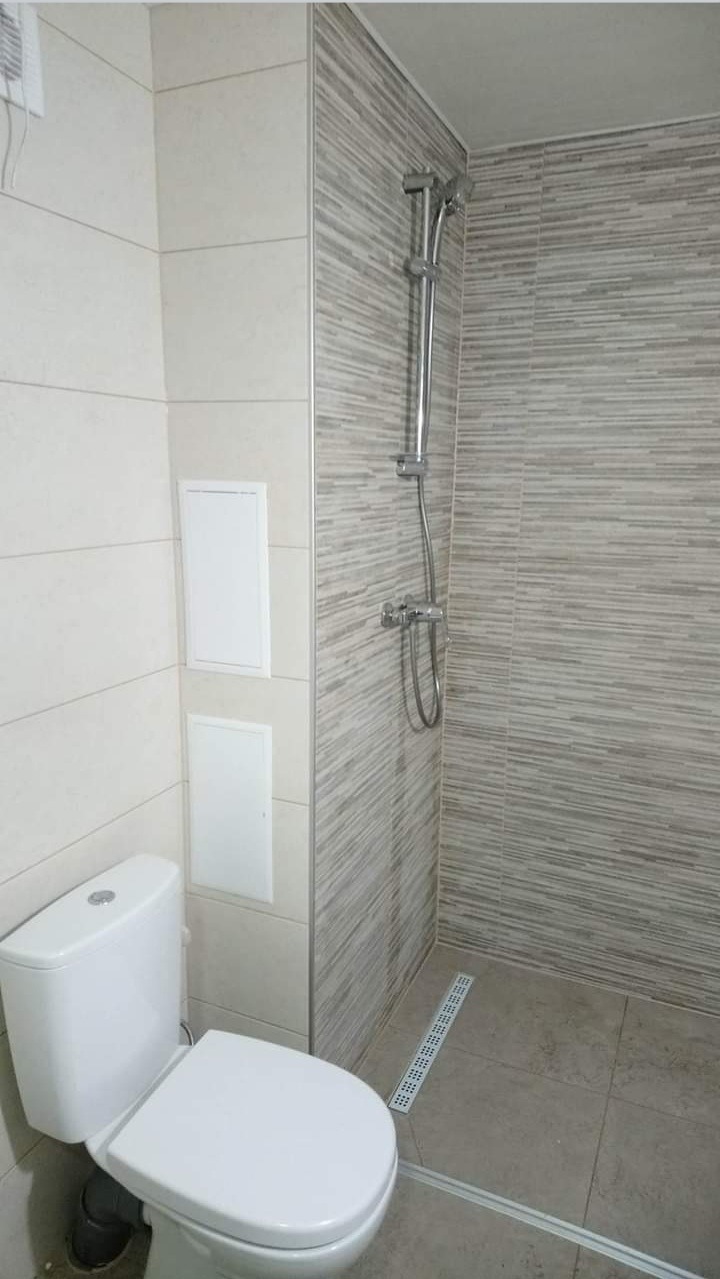 Nowoczesna łazienka z toaletą, kabiną prysznicową z deszczownicą i odpływem liniowym w podłodze, wykończona płytkami w odcieniach beżu i szarości z ukrytymi drzwiczkami rewizyjnymi.