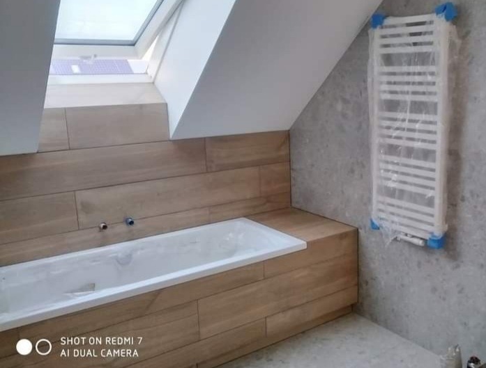 Łazienka na poddaszu w trakcie wykończenia: wanna obudowana drewnopodobnymi płytkami, grzejnik drabinkowy zabezpieczony folią, okno dachowe.