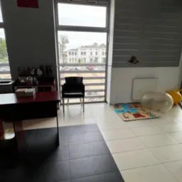 Gabinet z biurkiem, komputerem, krzesłem, zabawkami i matą edukacyjną, widok na budynek zza okna.