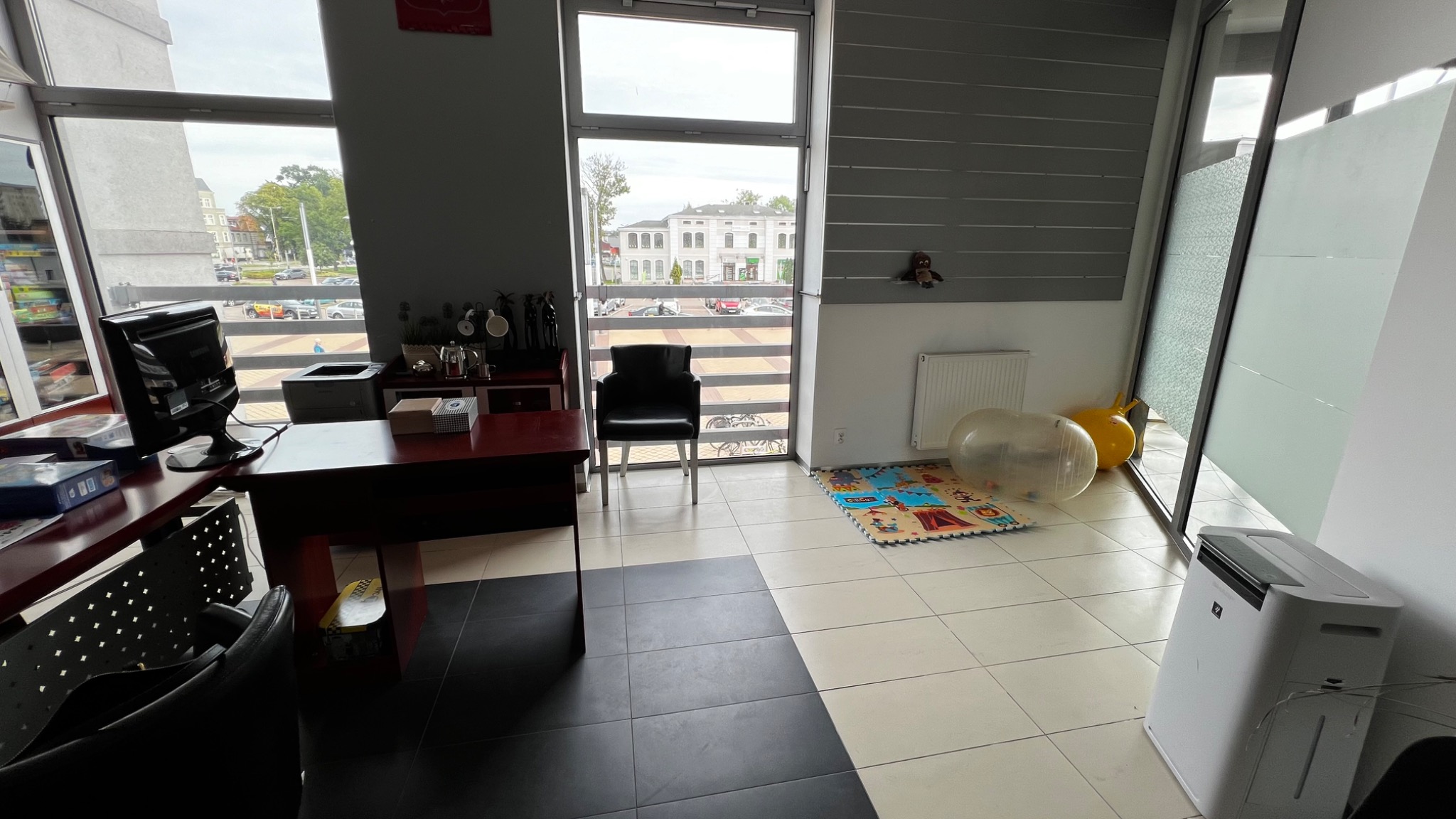 Gabinet z biurkiem, komputerem, krzesłem, zabawkami i matą edukacyjną, widok na budynek zza okna.