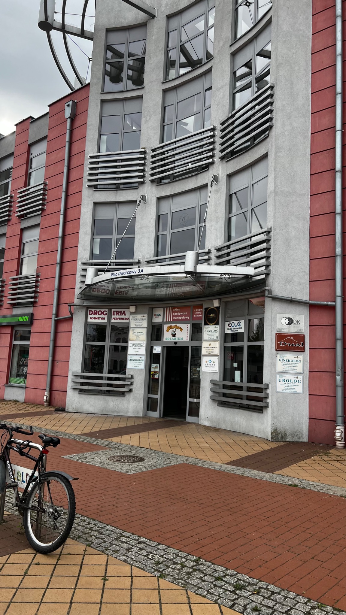 Wejście do budynku usługowego z licznymi tabliczkami informacyjnymi, w tym z napisem 'Logopeda', rower zaparkowany przed budynkiem.