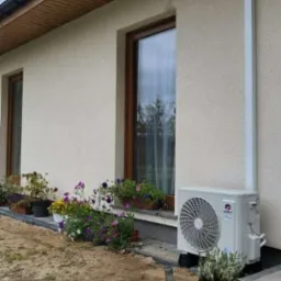 Zewnętrzna jednostka klimatyzacji zamontowana przy ścianie domu, z widoczną rurą odprowadzającą, otoczona kwiatami w doniczkach i wyłożonym kostką brukową chodnikiem.