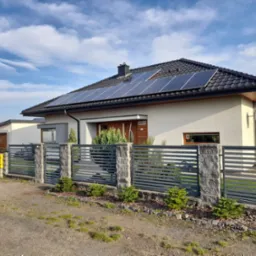 Nowoczesny dom jednorodzinny z panelami fotowoltaicznymi na dachu, ogrodzony metalowym płotem z elementami kamiennymi i żółtą skrzynką gazową przy bramie. Na dachu widoczny komin. Błękitne niebo...