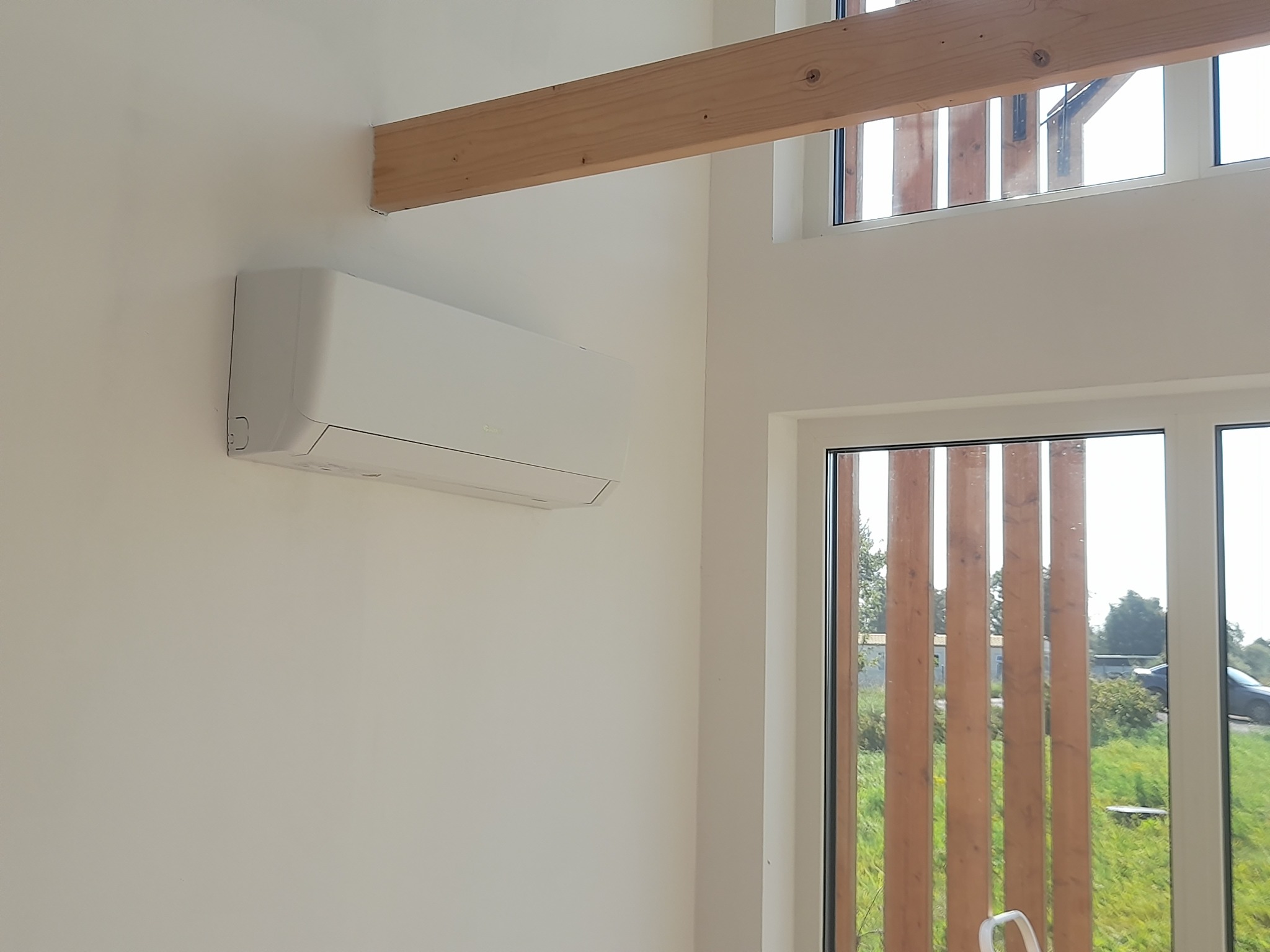 Biały klimatyzator ścienny w nowoczesnym wnętrzu z dużym oknem i drewnianą belką stropową.