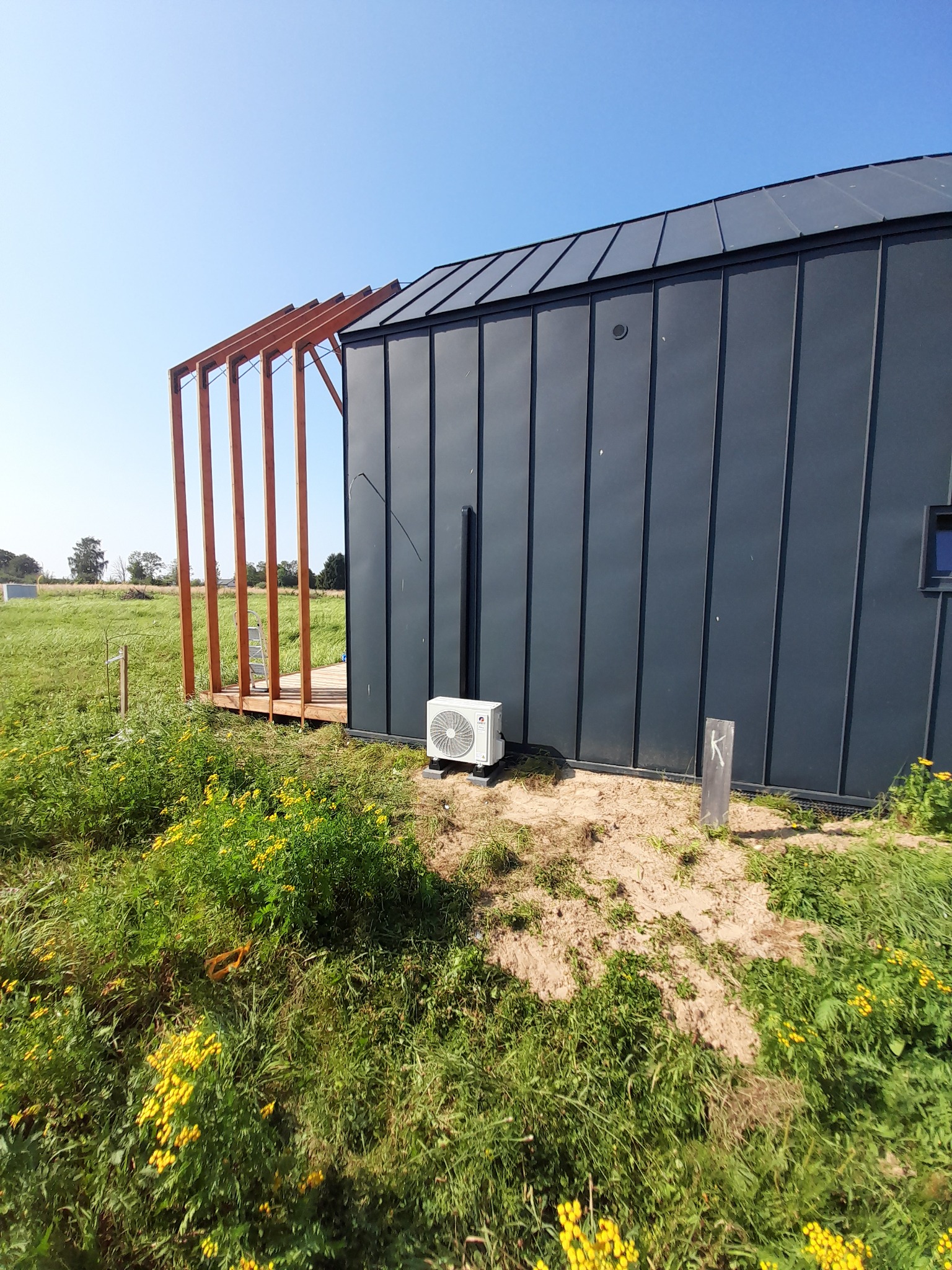 Nowoczesny dom z ciemnej blachy z klimatyzatorem na zewnątrz, drewniana konstrukcja tarasu w trakcie budowy, otoczony zielenią i żółtymi kwiatami.