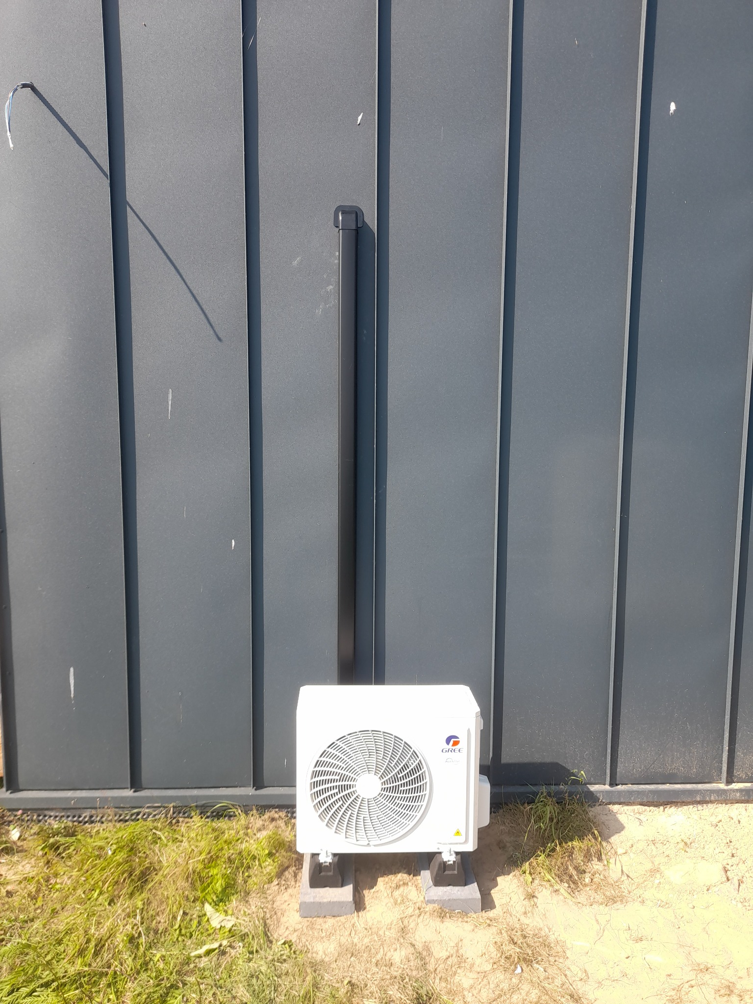 Biała jednostka zewnętrzna klimatyzatora marki Gree, zamontowana na betonowych podstawach przy ciemnoszarej, pionowej ścianie z blachy trapezowej, z widoczną rurą odprowadzającą skropliny.