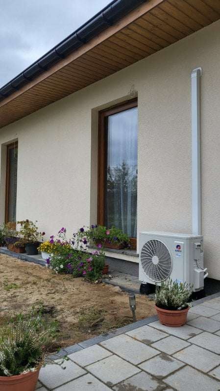 Zewnętrzna jednostka klimatyzacji zamontowana przy ścianie domu, z widoczną rurą odprowadzającą, otoczona kwiatami w doniczkach i wyłożonym kostką brukową chodnikiem.