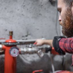 P.H.U Mateusz Majchrzak Hydro-Mat - Mężczyzna z brodą w czerwonej koszuli flanelowej reguluje zawór na czerwonym zbiorniku ciśnieniowym w instalacji grzewczej na tle szarej ściany.