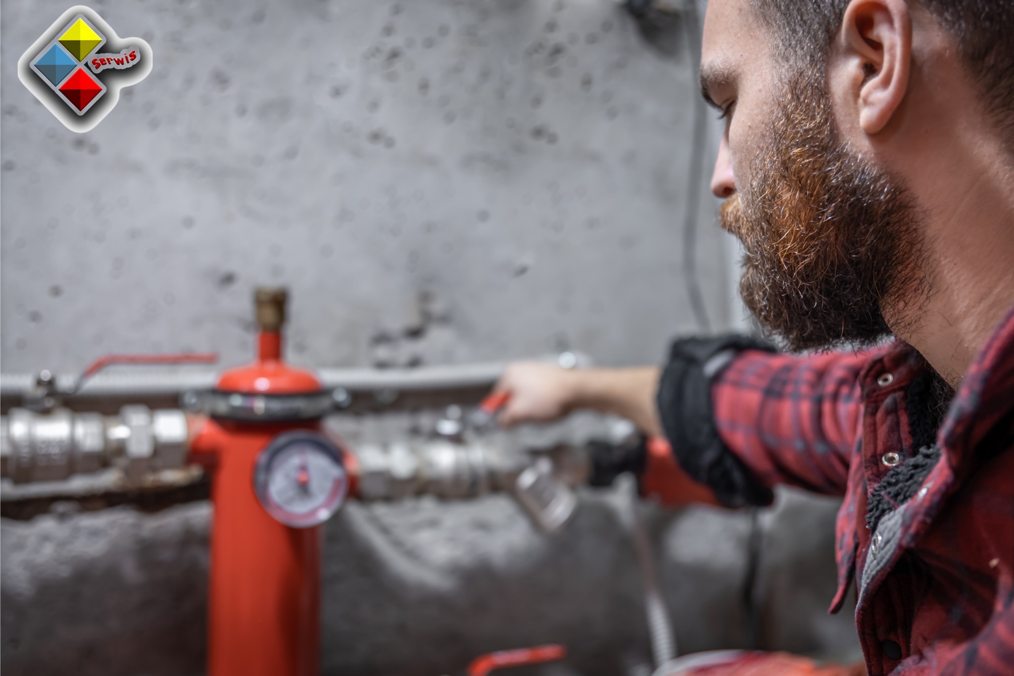 Mężczyzna z brodą w czerwonej koszuli flanelowej reguluje zawór na czerwonym zbiorniku ciśnieniowym w instalacji grzewczej na tle szarej ściany.