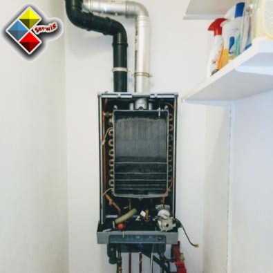 Otwarty piec gazowy z widocznymi elementami wewnętrznymi, rurami doprowadzającymi i odprowadzającymi, zamontowany na białej ścianie. Na górze po prawej widoczna biała półka z detergentami.