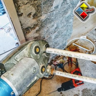 Zaciskanie złączki na rurze PEX/AL/PEXb narzędziem hydraulicznym, widoczna wiertarka udarowa i fragment ściany z surowego kamienia w tle.