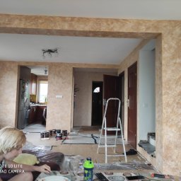 Perfectprint Malowanie agregatem - Salon w trakcie malowania dekoracyjnego z widoczną drabiną, puszkami farb i zabezpieczonymi powierzchniami; widoczny fragment osoby w lewym dolnym rogu.