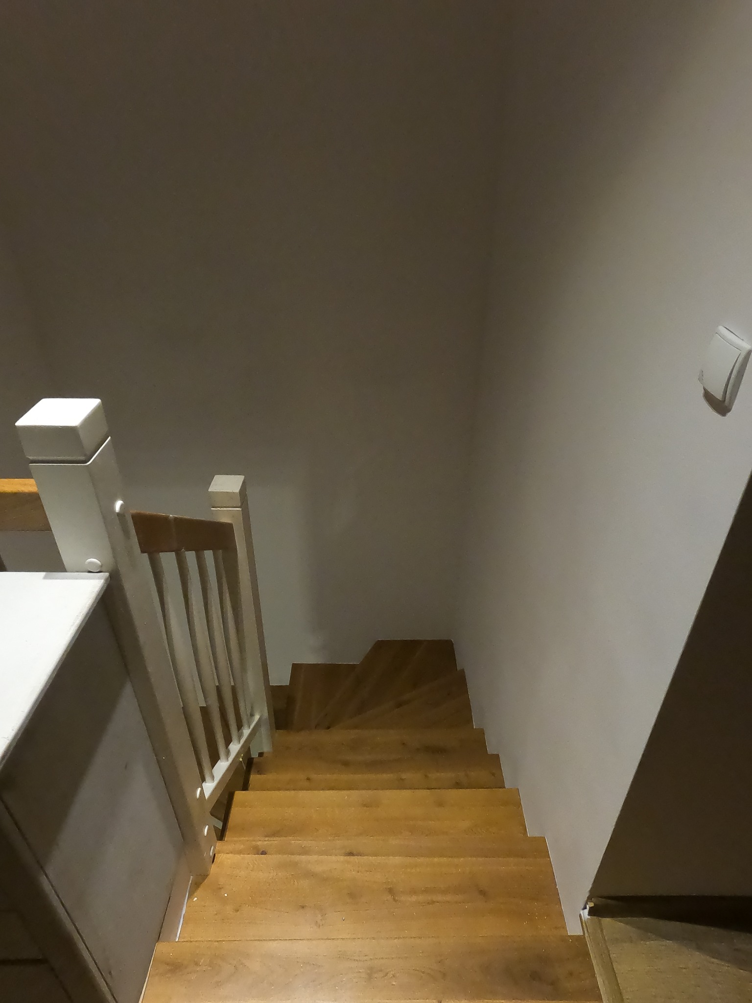 Drewniane schody w jasnym odcieniu drewna prowadzą w dół, widoczne białe balustrady po lewej stronie i biały włącznik światła na ścianie po prawej stronie.