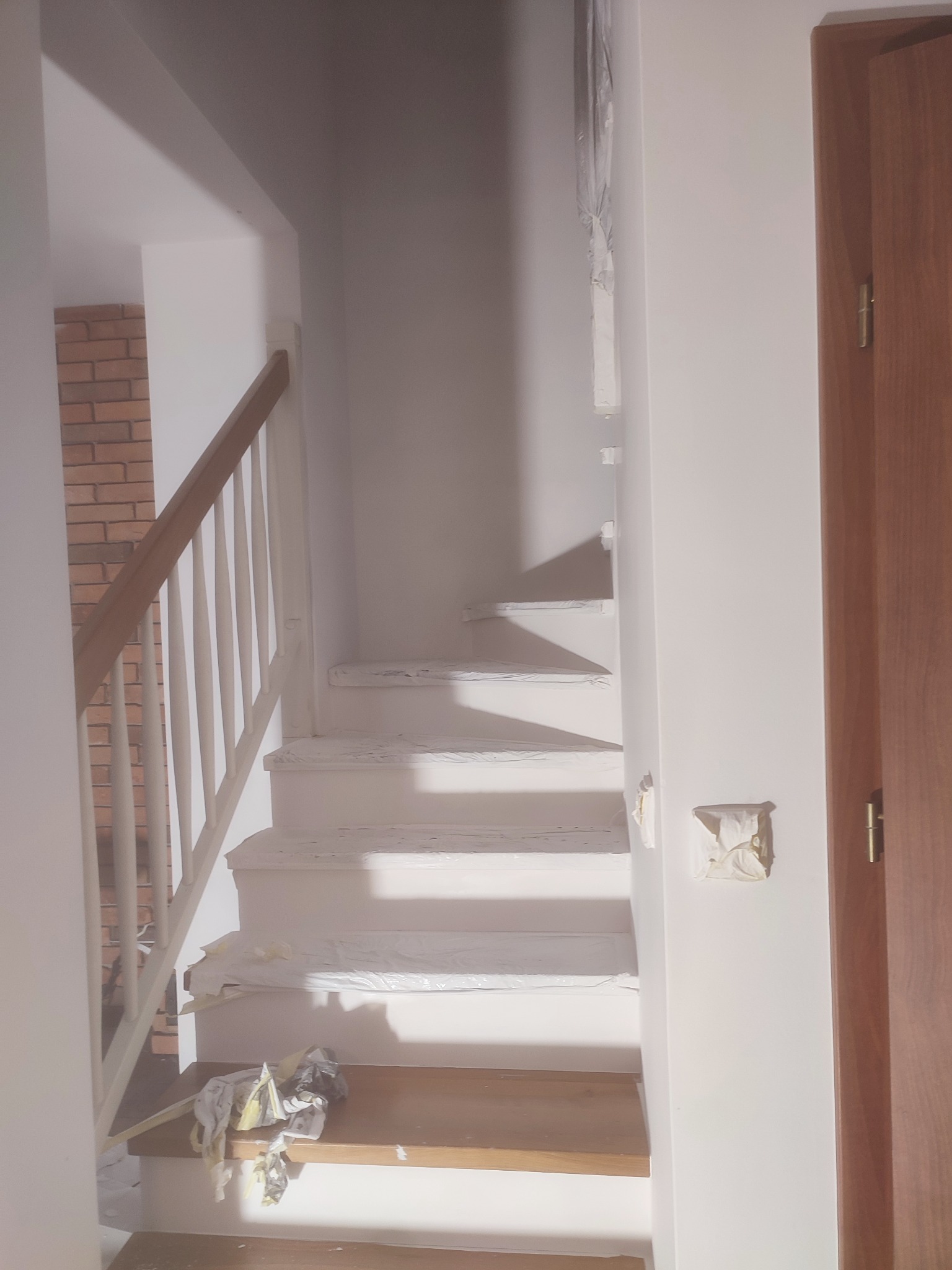 Schody w trakcie malowania na biało, zabezpieczone folią malarską, widoczny fragment ceglanej ściany w tle.