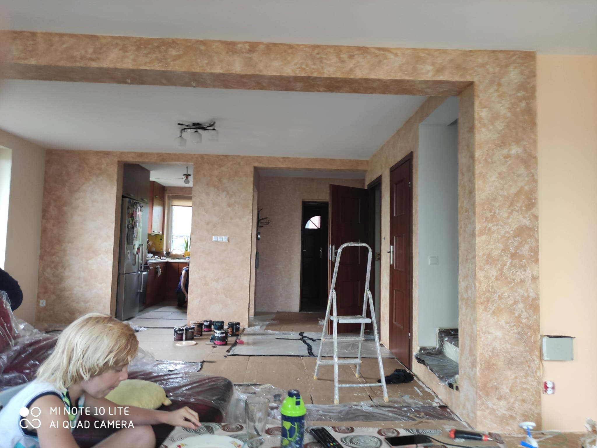 Salon w trakcie malowania dekoracyjnego z widoczną drabiną, puszkami farb i zabezpieczonymi powierzchniami; widoczny fragment osoby w lewym dolnym rogu.