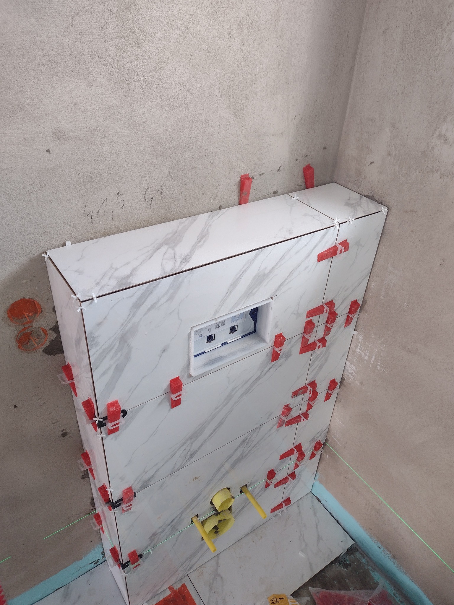 Montaż płytek imitujących marmur na ścianie z wnęką na stelaż WC, widoczne czerwone klipsy poziomujące i krzyżyki dystansowe, laserowy poziom wyznaczający linię, surowa ściana w tle.