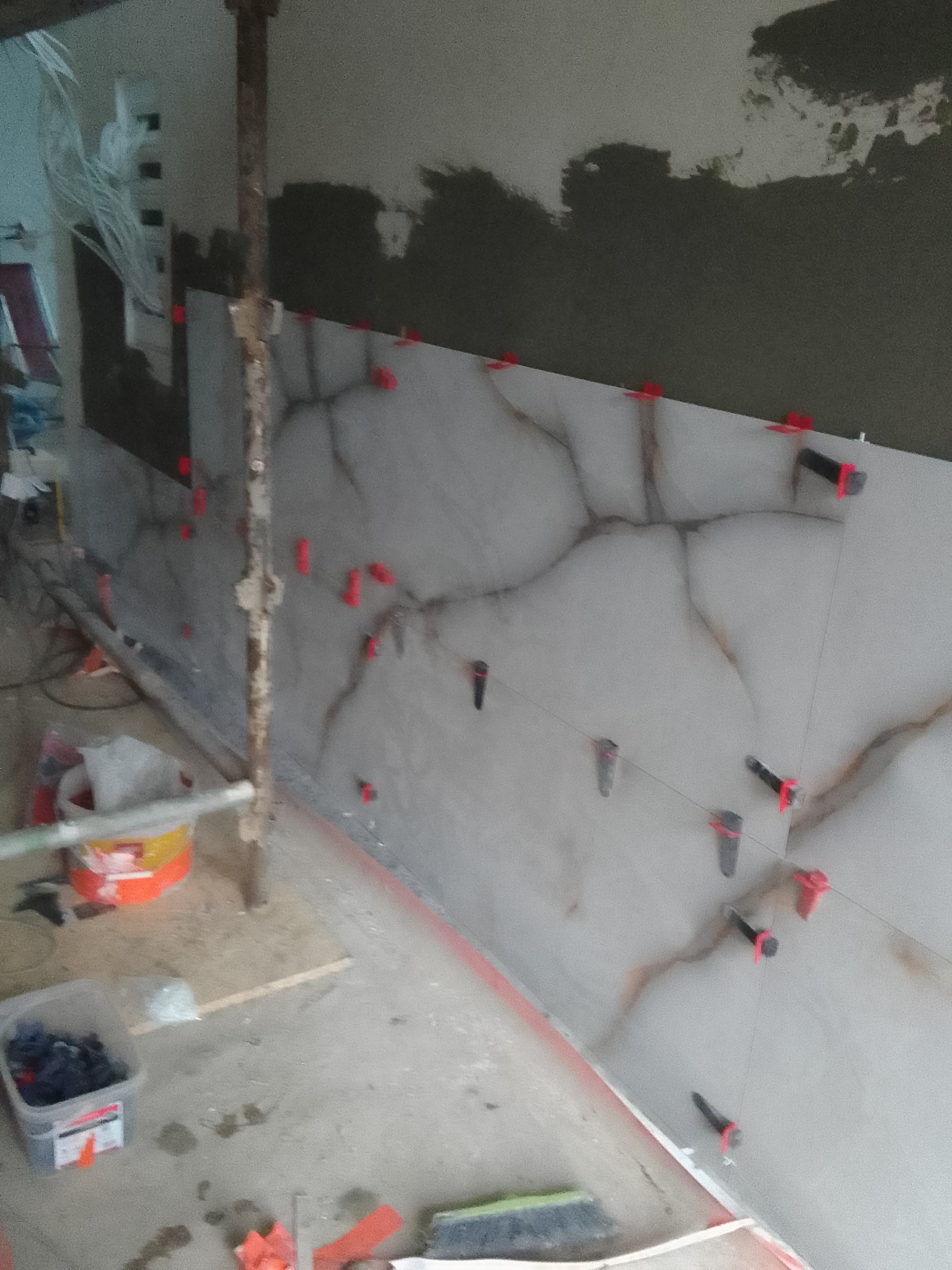 Ściana z ułożonymi płytkami imitującymi marmur, widoczne klipsy i kliny do poziomowania, ślady zaprawy na ścianie powyżej płytek, w tle widoczne narzędzia i materiały budowlane.