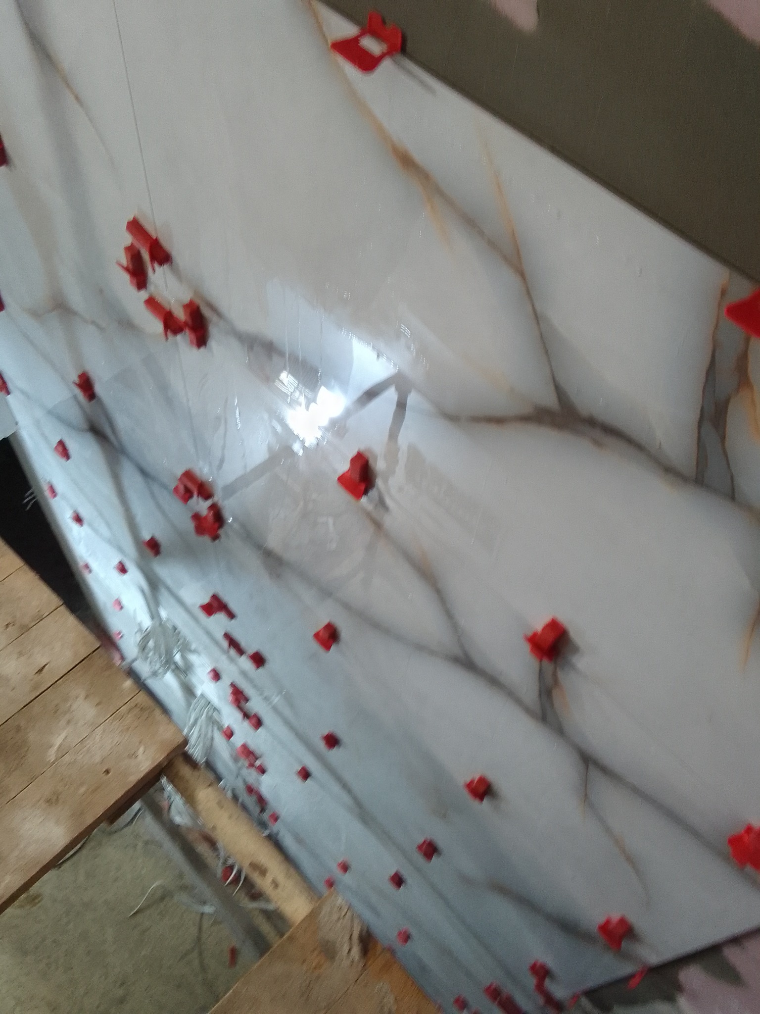 Układanie płytek imitujących marmur na ścianie z użyciem czerwonych klipsów poziomujących, widoczne drewniane elementy podłogi.