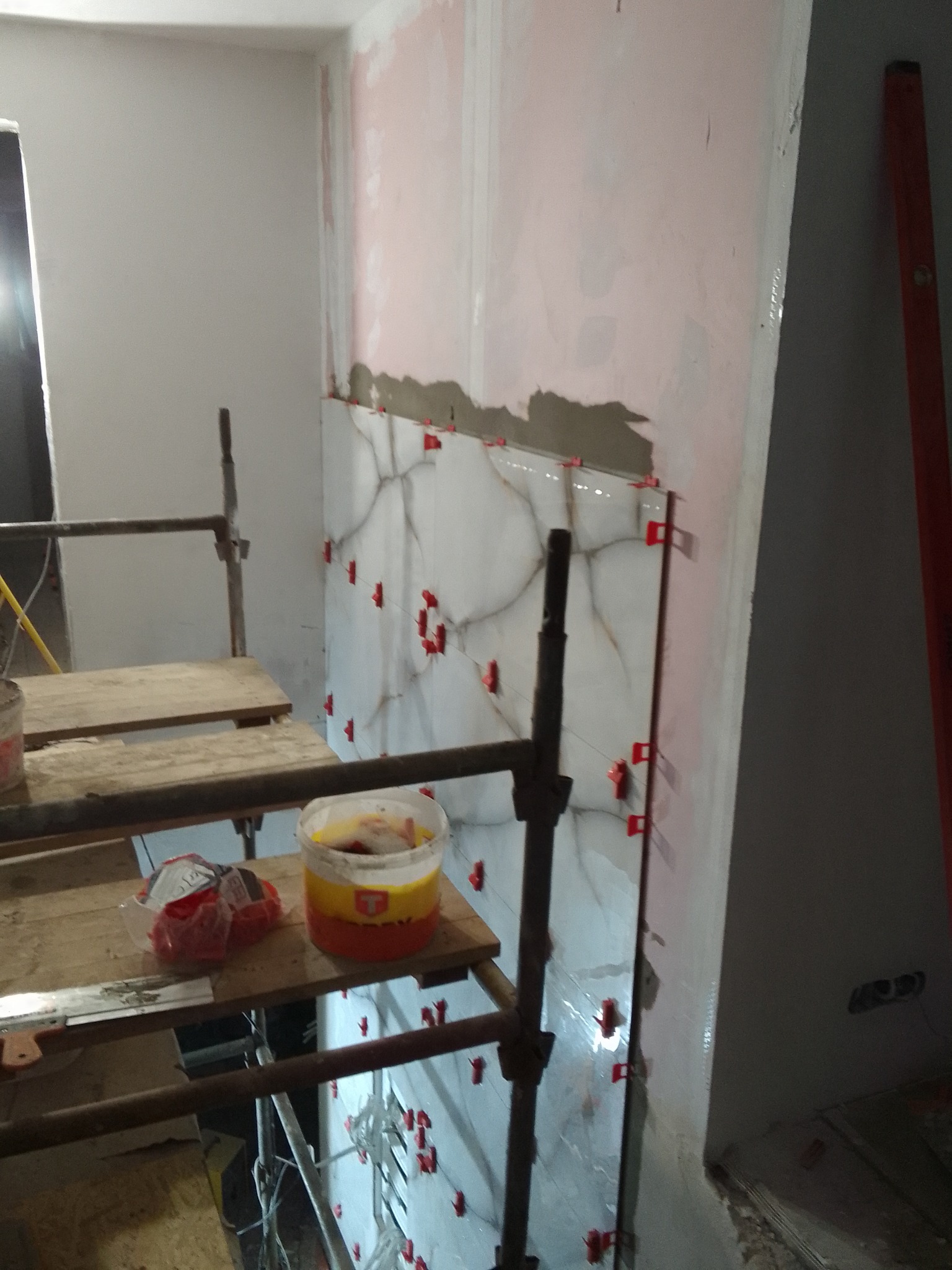Ściana w trakcie układania białych płytek imitujących marmur, z użyciem czerwonych krzyżyków dystansowych, widoczne rusztowanie i wiadro z zaprawą.