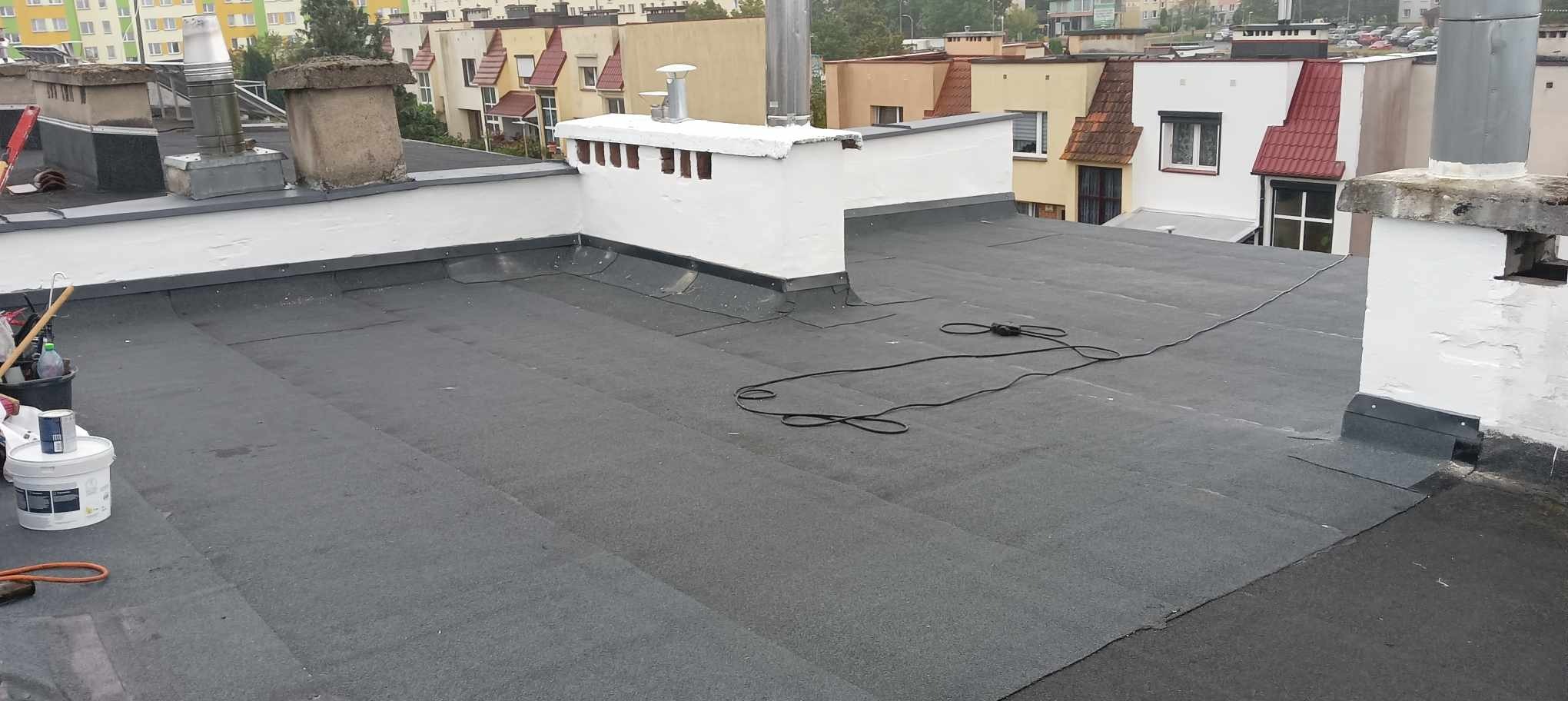 Płaski dach pokryty papą termozgrzewalną z widocznymi kominami i narzędziami pracy, w tle zabudowa miejska.
