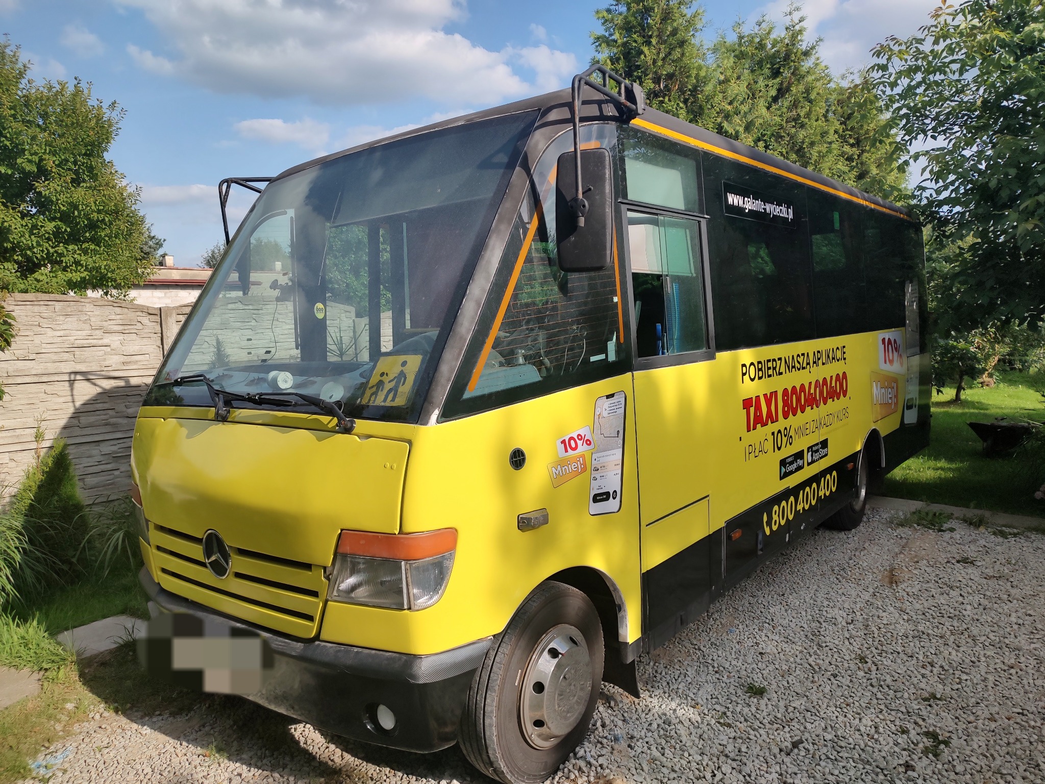 Żółto-czarny bus Mercedes-Benz z reklamą aplikacji taxi, zaparkowany na żwirowym podłożu, z widocznym logo firmy i numerem telefonu.