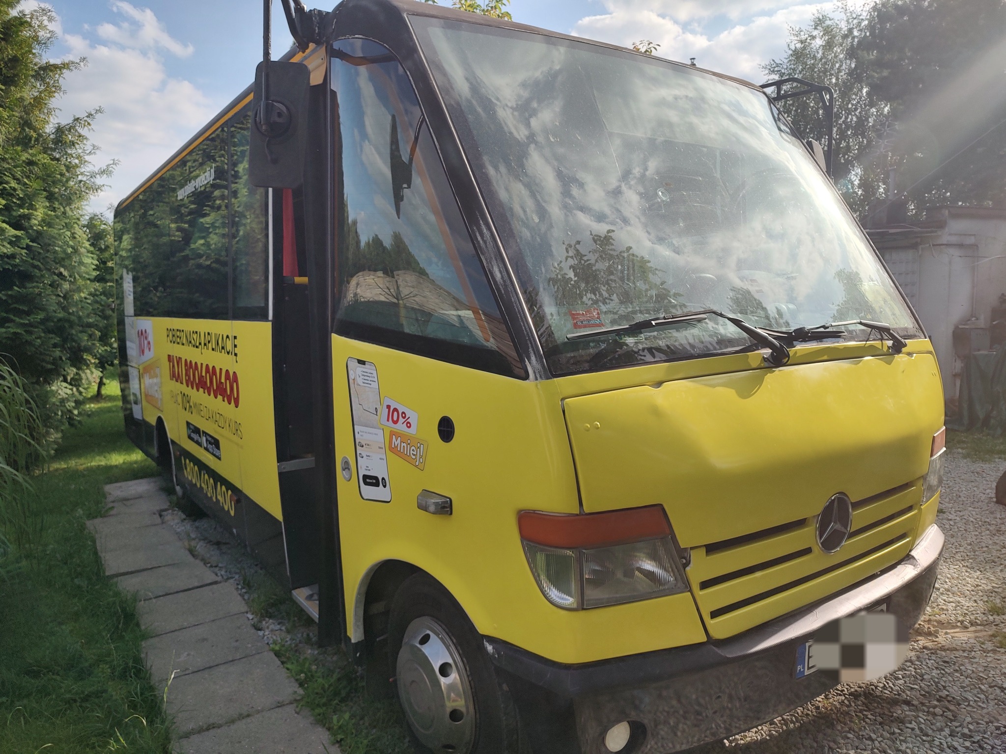 Żółty bus Mercedes z reklamą aplikacji mobilnej do zamawiania taksówek, zaparkowany na zewnątrz, widok z przodu i boku.