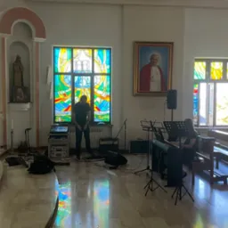 Przygotowania do występu muzycznego w kościele: mikser, keyboard, wzmacniacz, mikrofony i nuty rozstawione przed kolorowymi witrażami, portret papieża w tle.