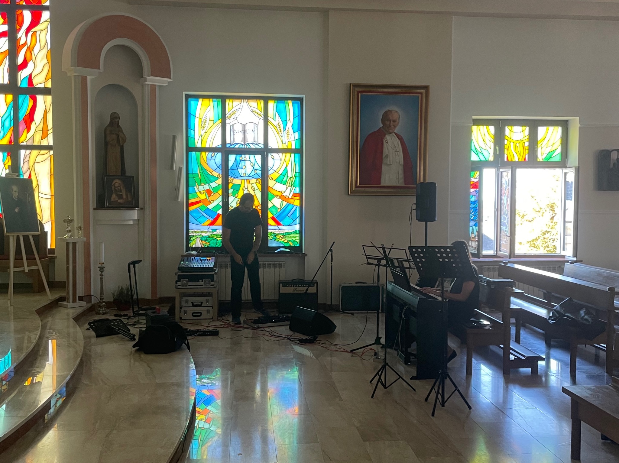 Przygotowania do występu muzycznego w kościele: mikser, keyboard, wzmacniacz, mikrofony i nuty rozstawione przed kolorowymi witrażami, portret papieża w tle.