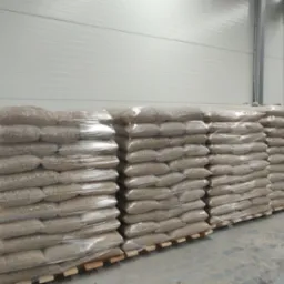 Magazynowane worki pelletu drzewnego ułożone na paletach, owinięte folią stretch, na tle białej, metalowej ściany hali.