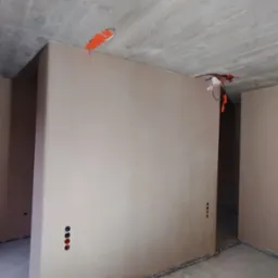 Wnętrze w stanie deweloperskim po nałożeniu gładzi gipsowej na ściany, widoczne punkty elektryczne zabezpieczone pomarańczową taśmą, surowy betonowy sufit.