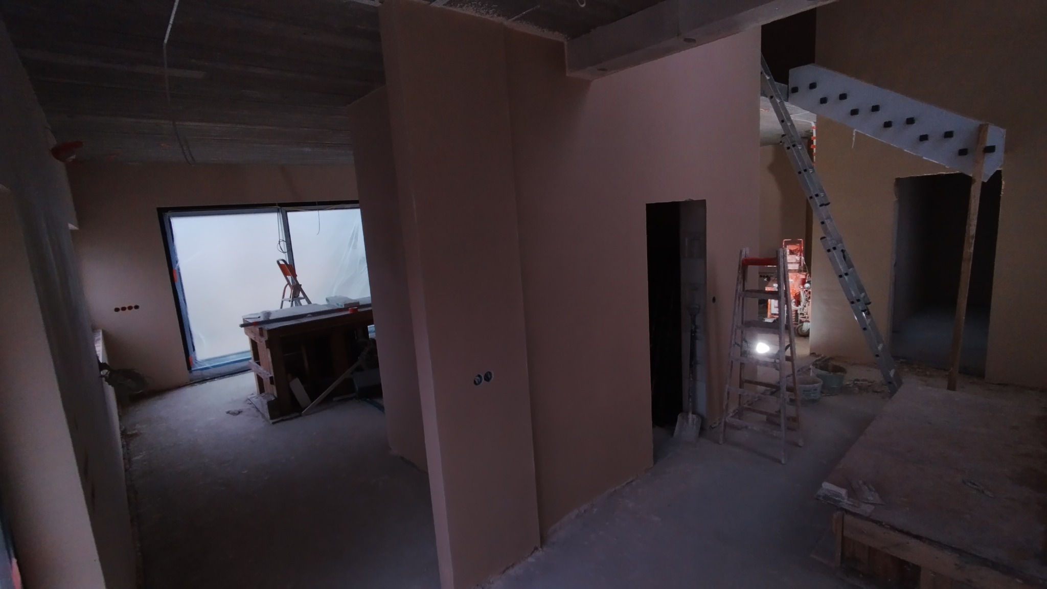 Wnętrze w trakcie tynkowania maszynowego: gładkie, jasnoróżowe ściany, widoczne puszki elektryczne, drabiny i narzędzia budowlane w tle, surowy betonowy sufit.
