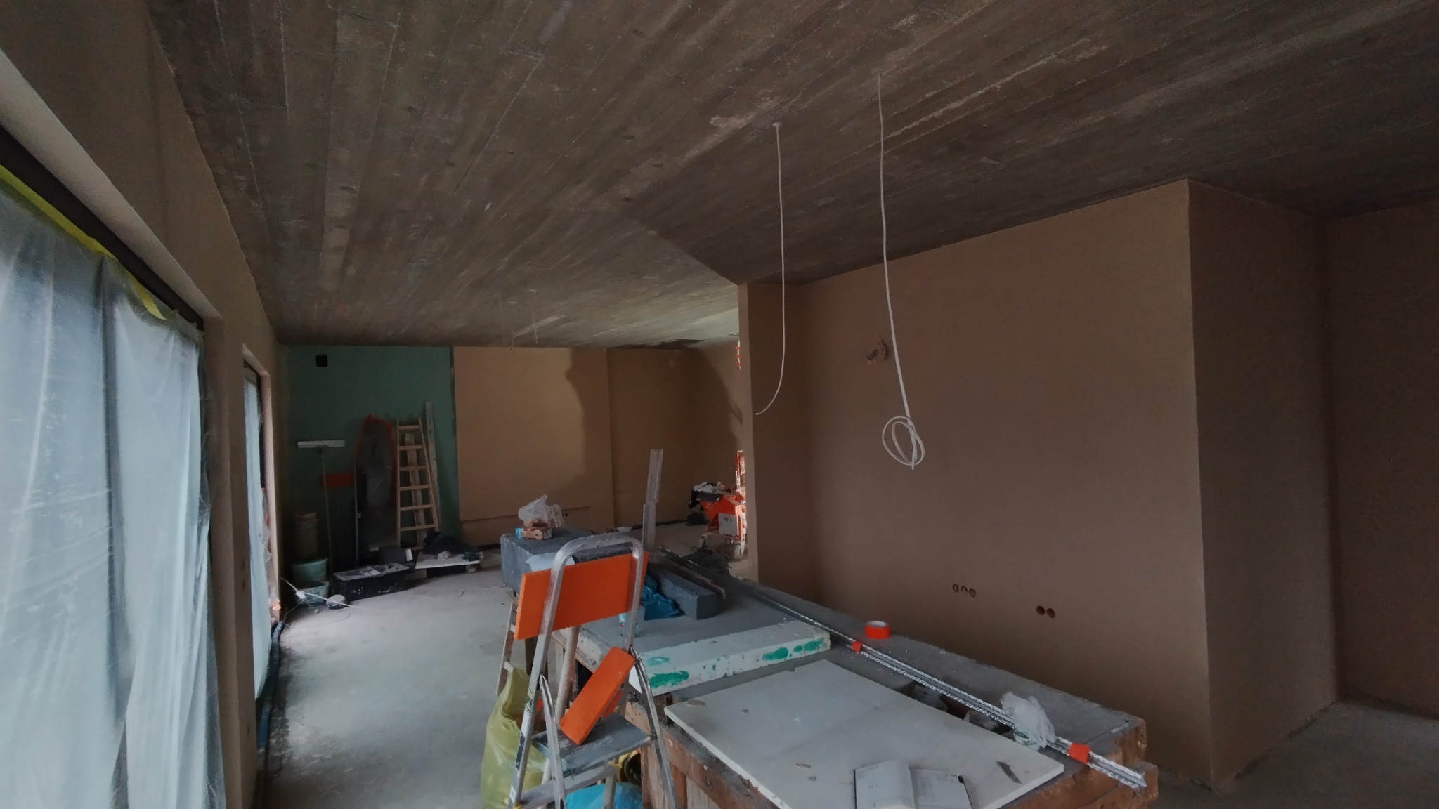 Wnętrze w trakcie prac wykończeniowych: ściany pokryte świeżym tynkiem, widoczne kable elektryczne zwisające z surowego stropu, drabina i materiały budowlane w tle.