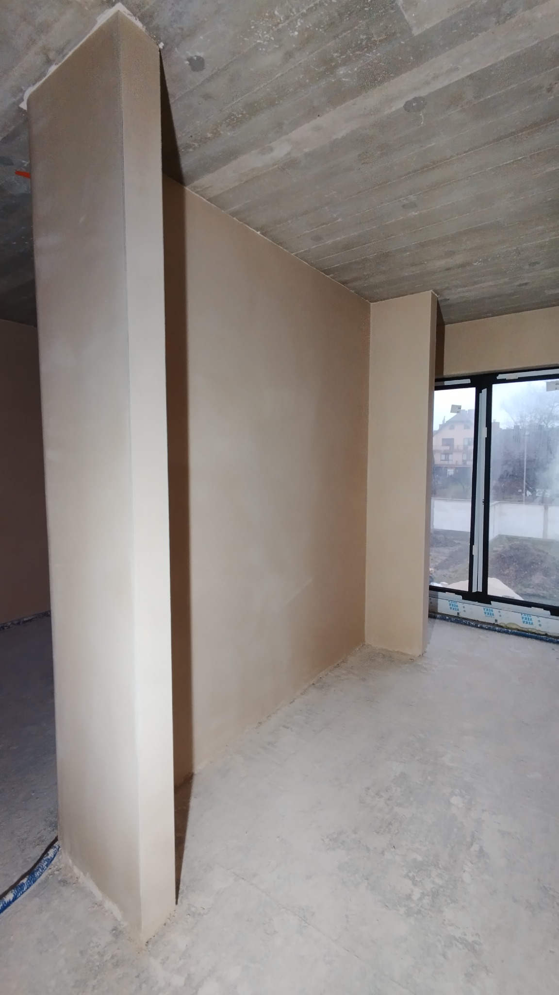 Świeżo otynkowane ściany i filar w surowym wnętrzu, widok na okno z zewnątrz na częściowo zagospodarowany teren.