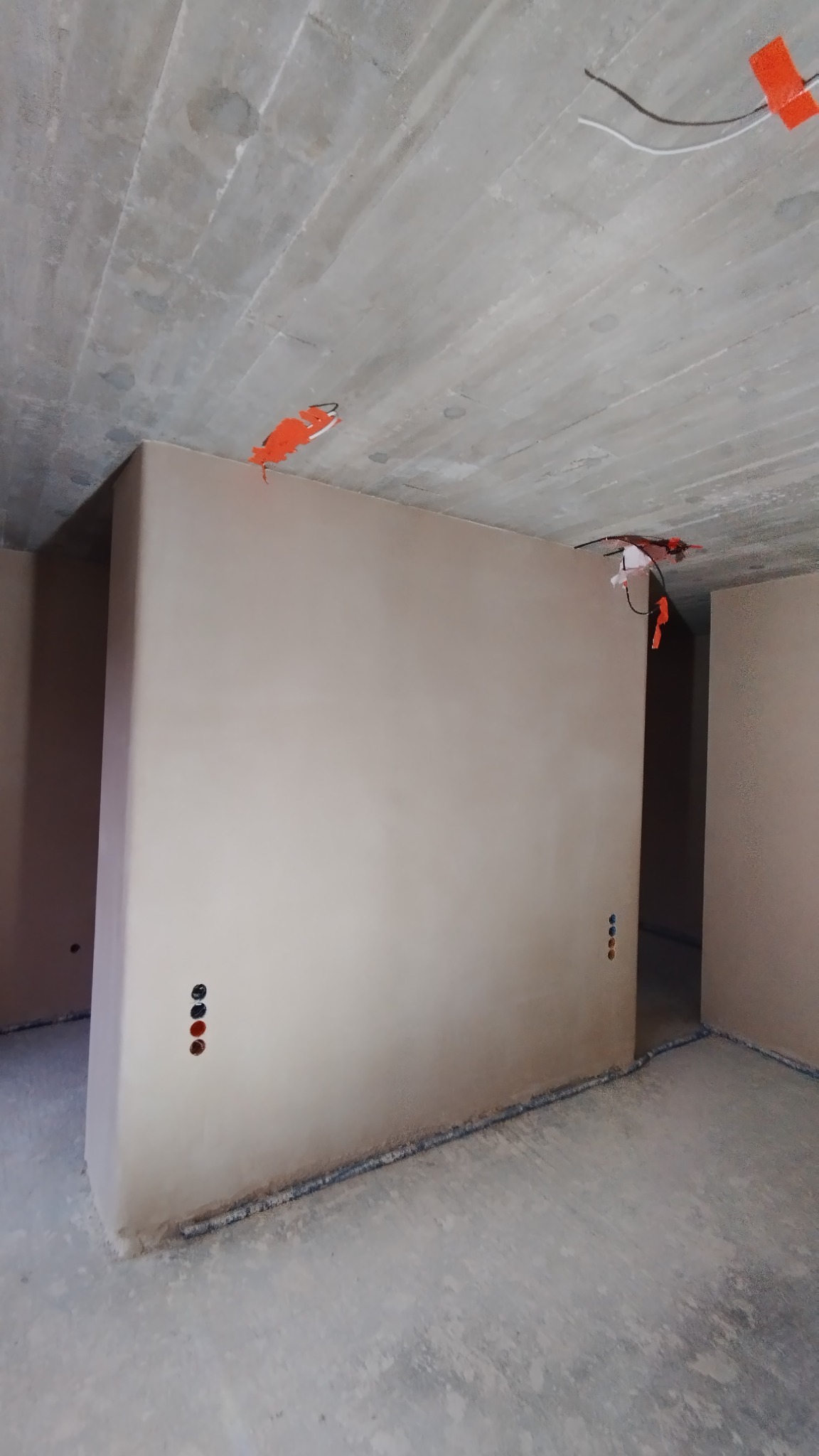 Wnętrze w stanie deweloperskim po nałożeniu gładzi gipsowej na ściany, widoczne punkty elektryczne zabezpieczone pomarańczową taśmą, surowy betonowy sufit.