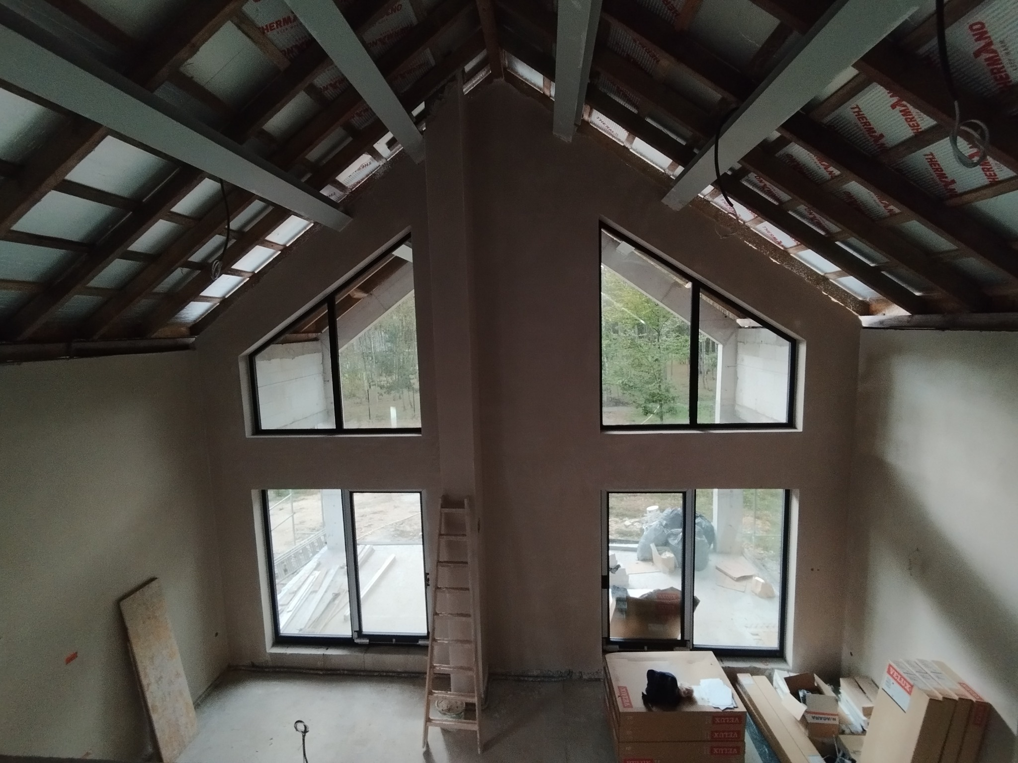 Wnętrze domu w trakcie wykańczania: ściany po nałożeniu gładzi, widoczne okna z czarnymi ramami, drewniana konstrukcja dachu z izolacją, drabina oparta o ścianę, kartony i narzędzia na podłodze.