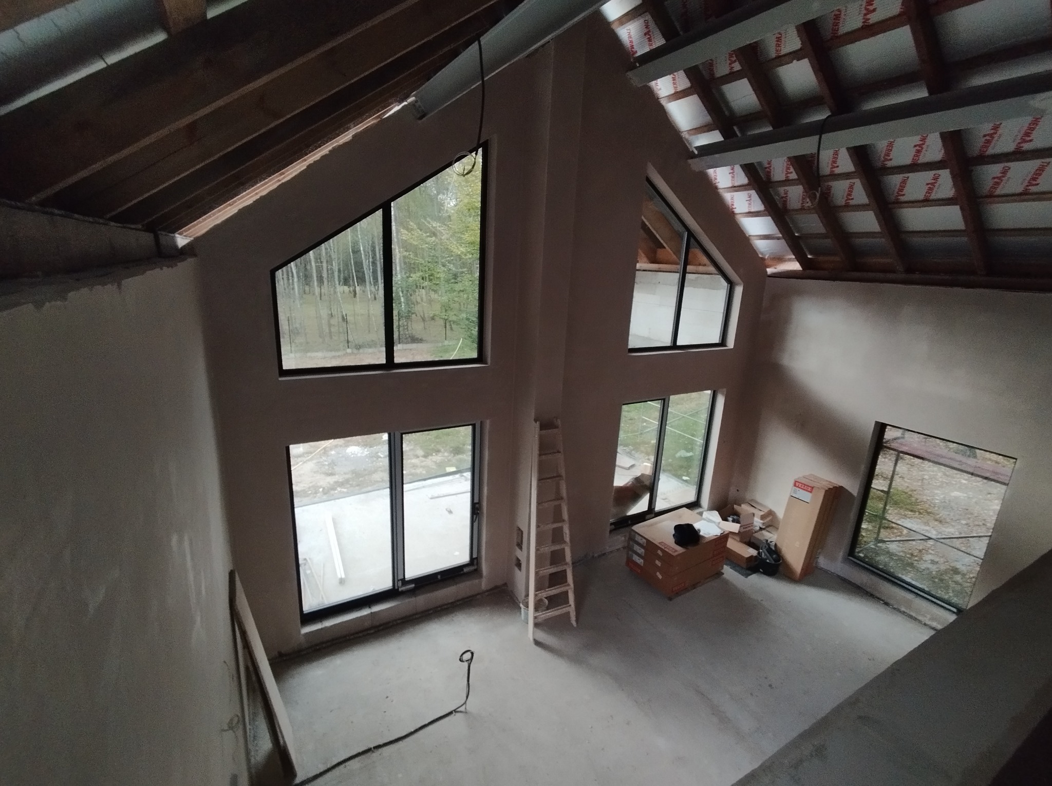 Surowe wnętrze domu w trakcie wykańczania, widoczne świeżo nałożone, gładkie ściany, drewniane krokwie dachu z izolacją, okna z czarnymi ramami i drabina oparta o ścianę.